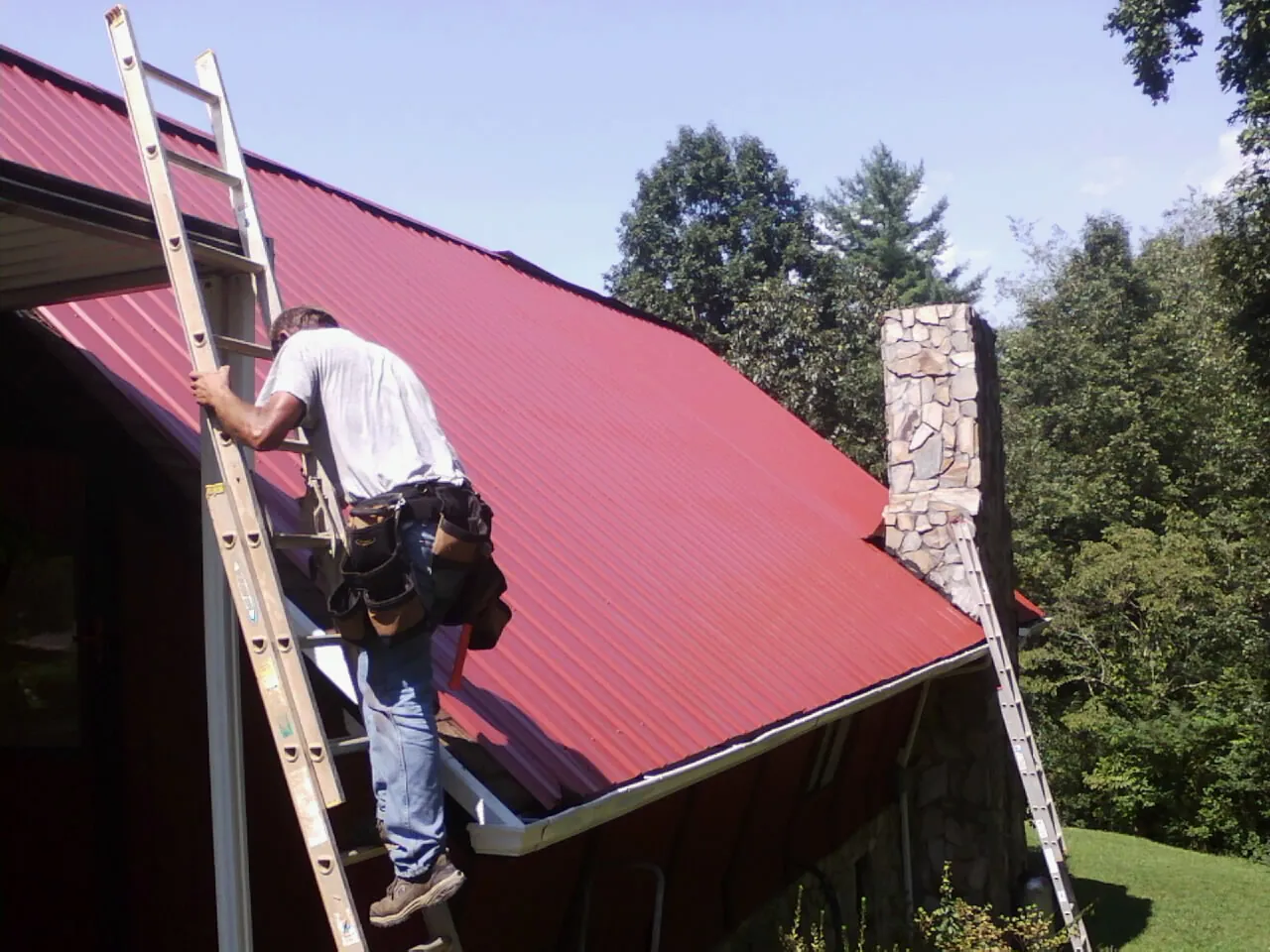 Roofer on a ladder beside a red metal roof and stone chimney — Soffit & Fascia Repair by Roofing Brothers in La Plata